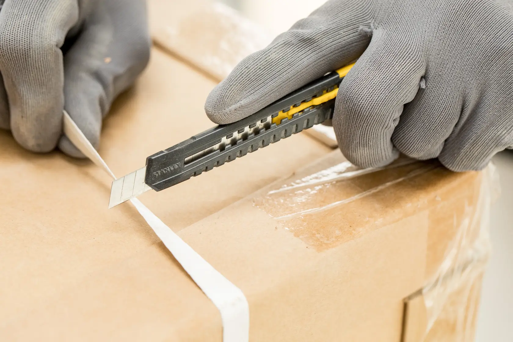 STANLEY 0-10-150 Universalmesser schneidet weißen Gurt auf Kartonbox, in der Hand, Nahaufnahme.