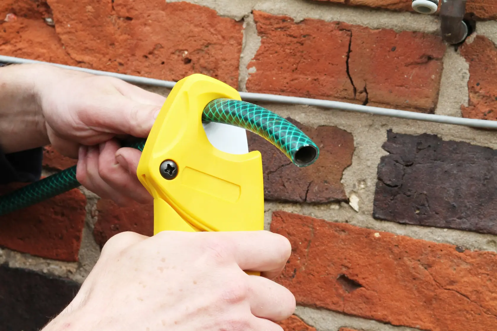 Foto des STANLEY 0-70-450 Schlauchschneiders beim Schneiden eines grünen Schlauchs an Ziegelwand.
