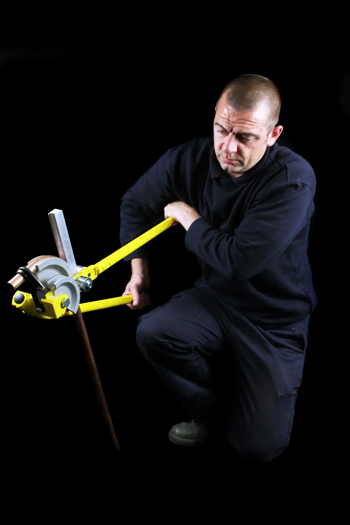 STANLEY 0-70-452 pipe bender in use bending a copper pipe, person kneeling, black background.