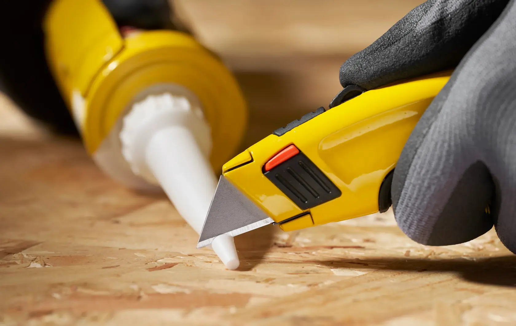 STANLEY 1-11-700 utility knife close-up cutting caulking tube nozzle on wood surface, photo from side angle.