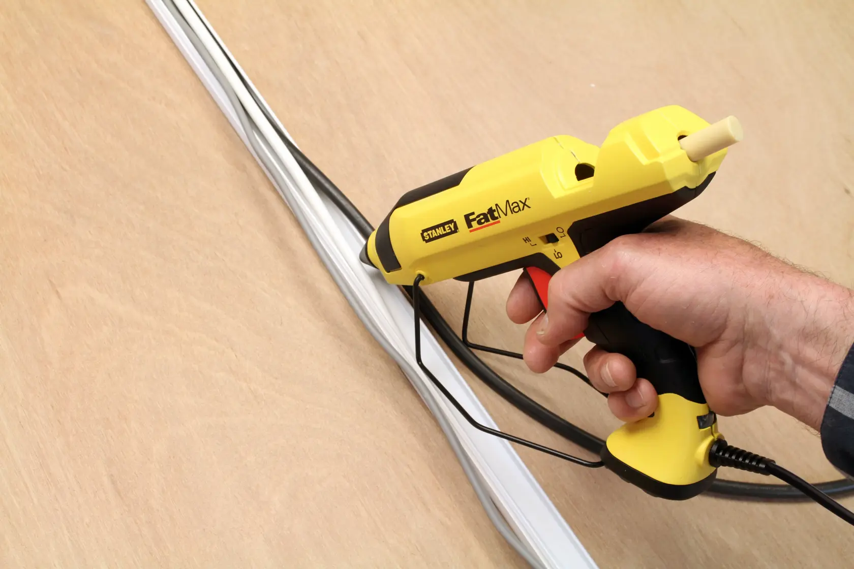 Stanley FMHT6-70418 glue gun held in hand, yellow and black, applying adhesive to wires on wood surface.