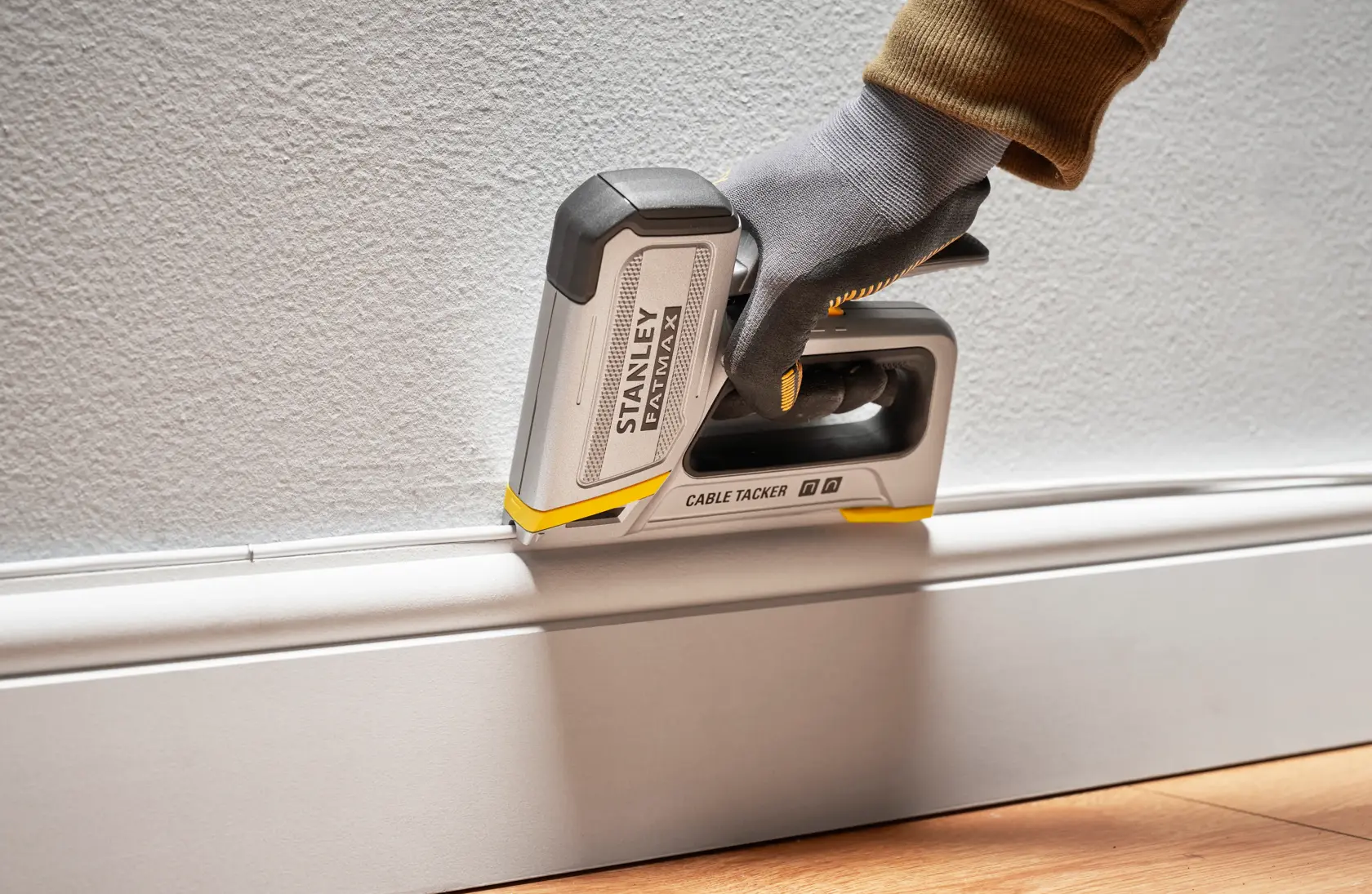 Stanley FMHT70100-0 cable tacker in use along baseboard, shown held by a gloved hand against wall.