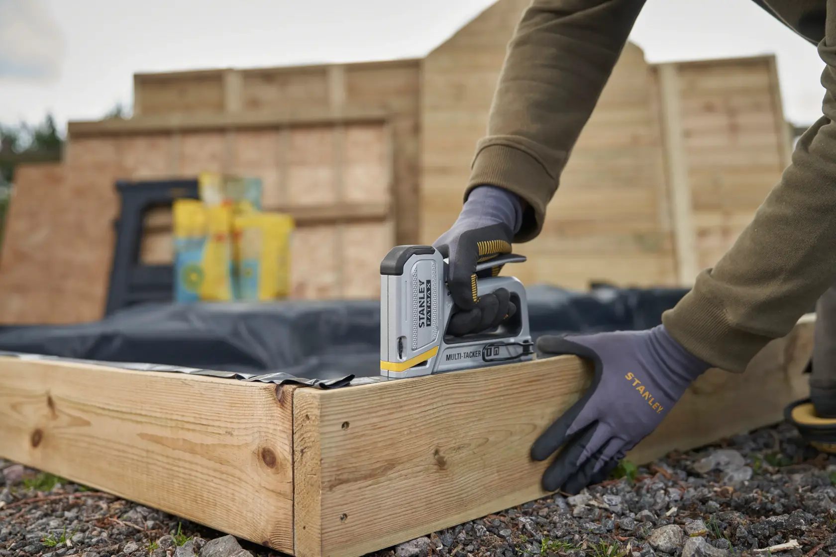 Photo of STANLEY FMHT70250-0 staple gun fastening black sheet to wooden frame outdoors, person wearing STANLEY gloves.