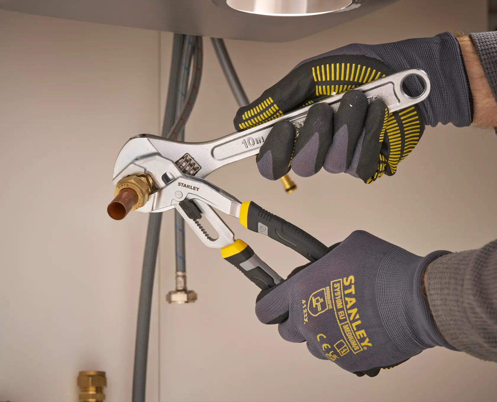 STANLEY FMHT75502-0 adjustable wrench in use, held by gloved hands tightening a pipe fitting under a sink.