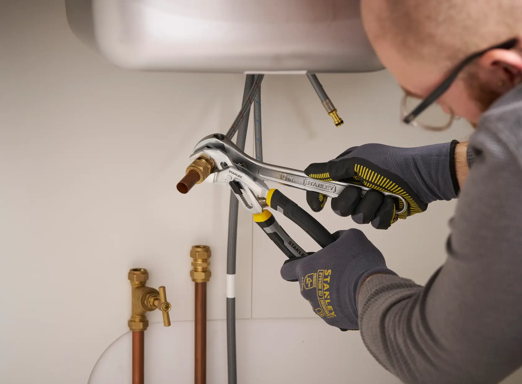 STANLEY FMHT75508-0 adjustable pliers in use under sink on plumbing pipes, hands with STANLEY gloves.