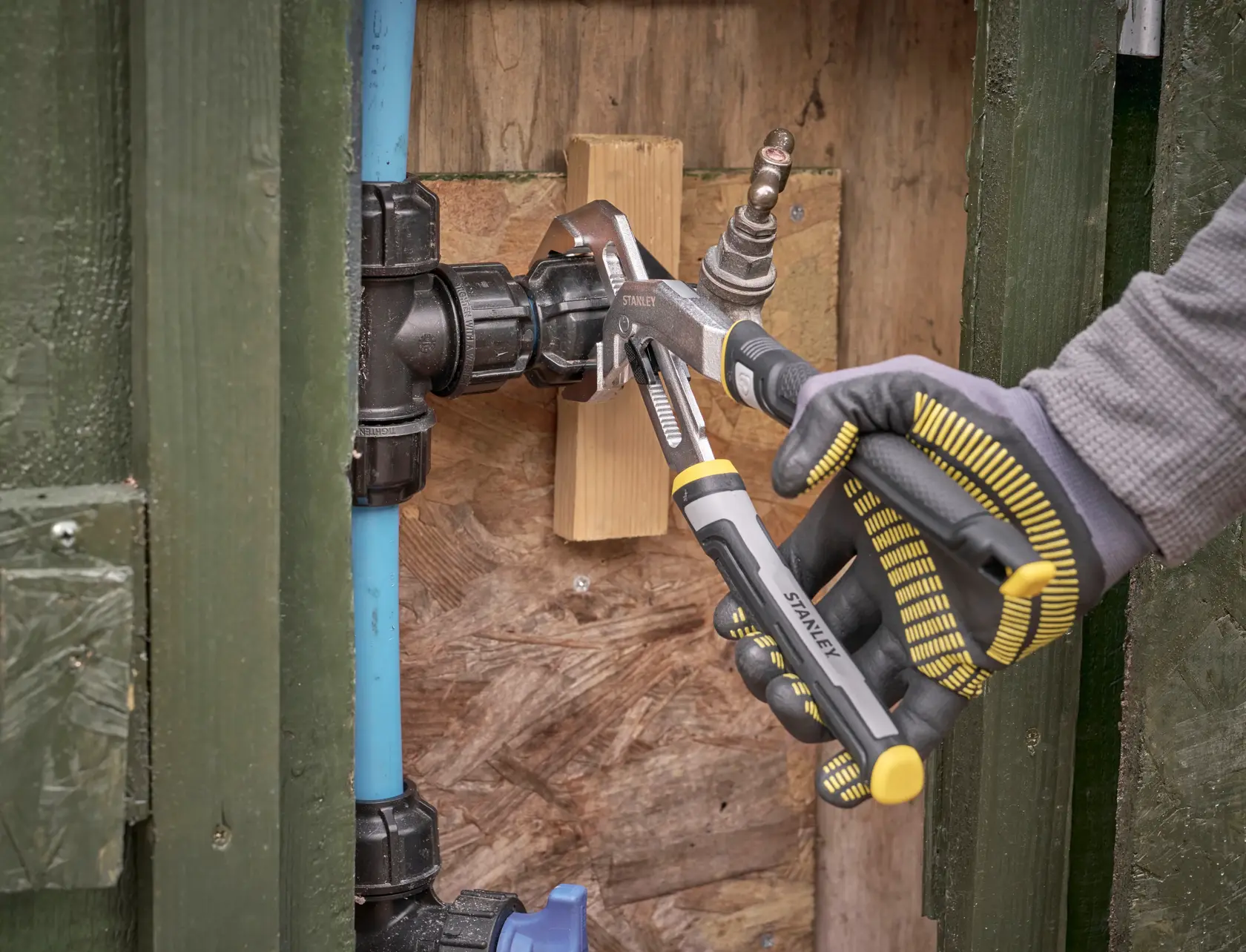 STANLEY FMHT75512-0 water pump pliers gripping a plumbing fitting, shown in use with gloved hand outdoors.