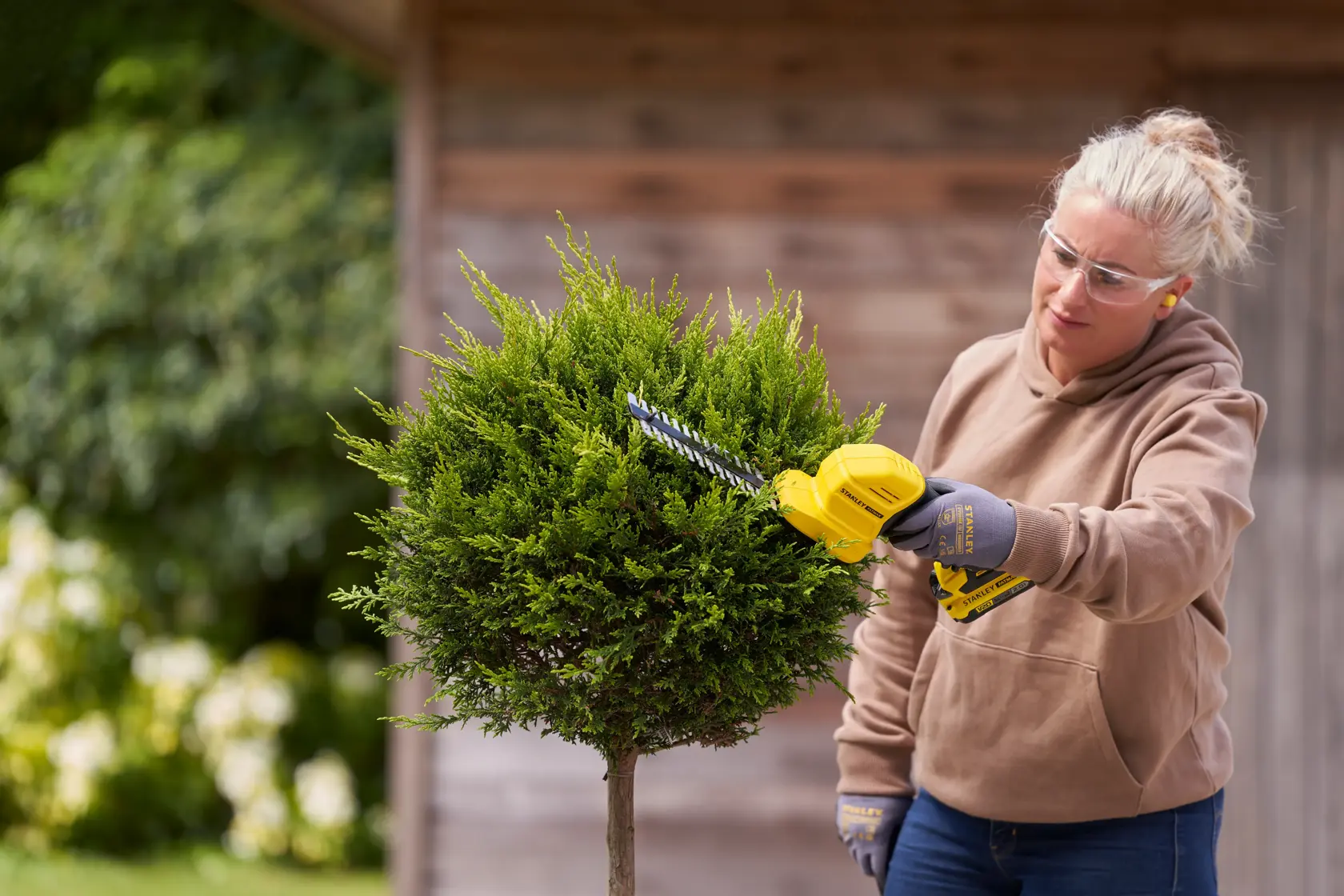 Photo of STANLEY STCSS20D1 hedge trimmer in use, trimming a green bush outdoors.