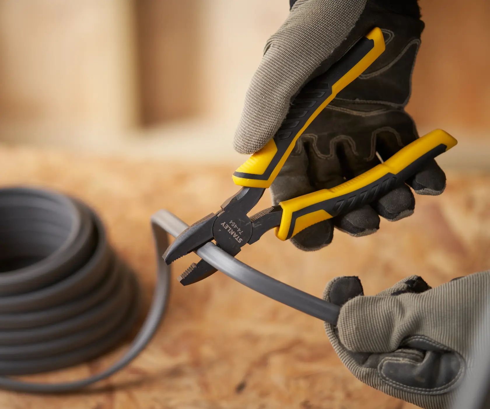 STANLEY STHT0-74454 pliers with yellow and black handle cutting thick gray wire, close-up, worn by gloved hands.