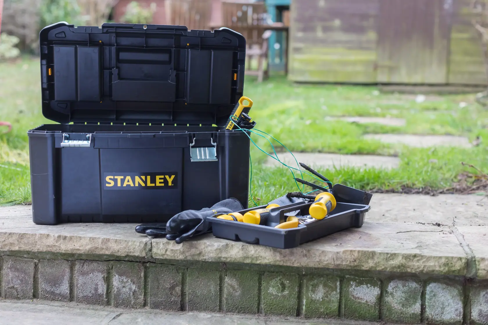 STANLEY STST1-75521 schwarze Werkzeugbox offen mit gelben Werkzeugen und Handschuhen auf einer Steinstufe.